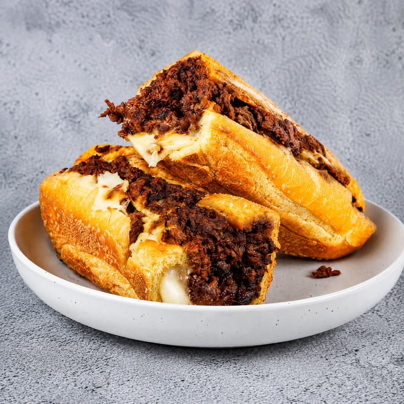 Two halves of a cheesy steak sandwich on a white plate against a gray background.