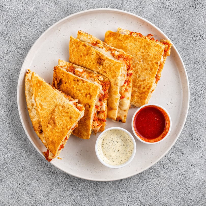 Golden-brown quesadilla slices filled with cheese and chicken, served with red and white dipping sauces.